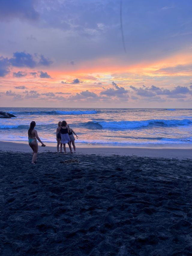 Un voyage de groupe WeRoad de femmes debout sur une plage de sable noir au bord de l'océan pendant un coucher de soleil coloré.