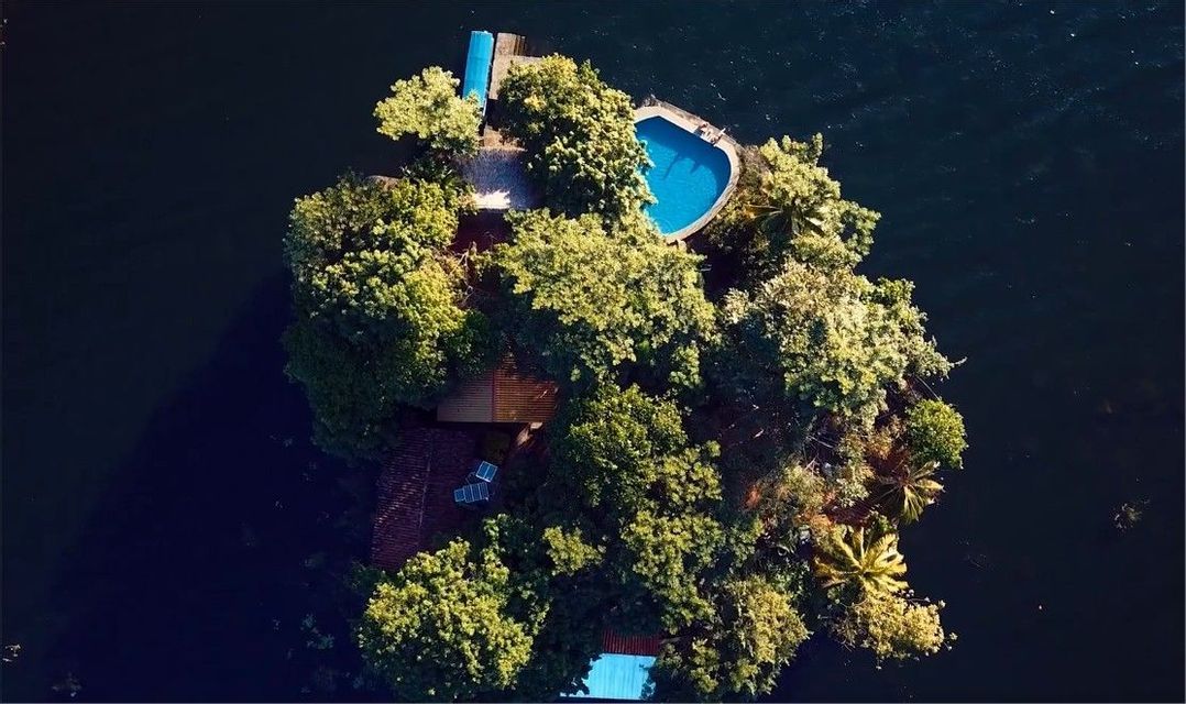 Vue aérienne d'une petite île verdoyante et luxuriante, dotée d'une piscine bleue et de plusieurs maisons, le tout entouré d'une eau sombre.