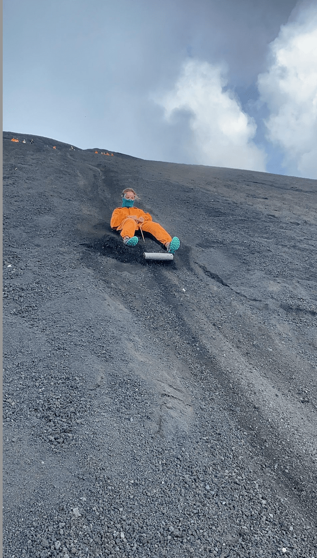 Une personne en combinaison de protection orange dévale une pente raide de cendres volcaniques sur une planche lors d'un voyage de groupe WeRoad.