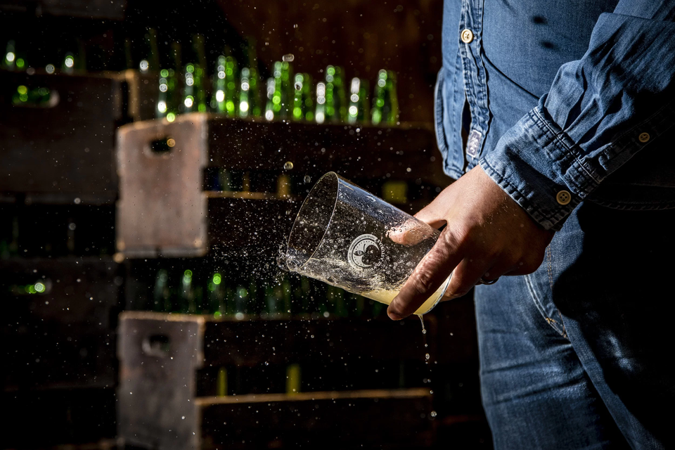 Una persona con camisa vaquera sostiene un vaso, salpicando una bebida amarilla dentro de una bodega oscura con botellas al fondo.