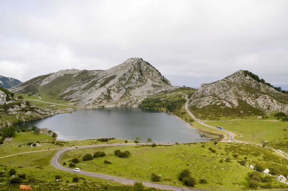 Un lago de montaña, rodeado de verdes colinas y picos rocosos, con carreteras para vehículos que serpentean a lo largo de su orilla.