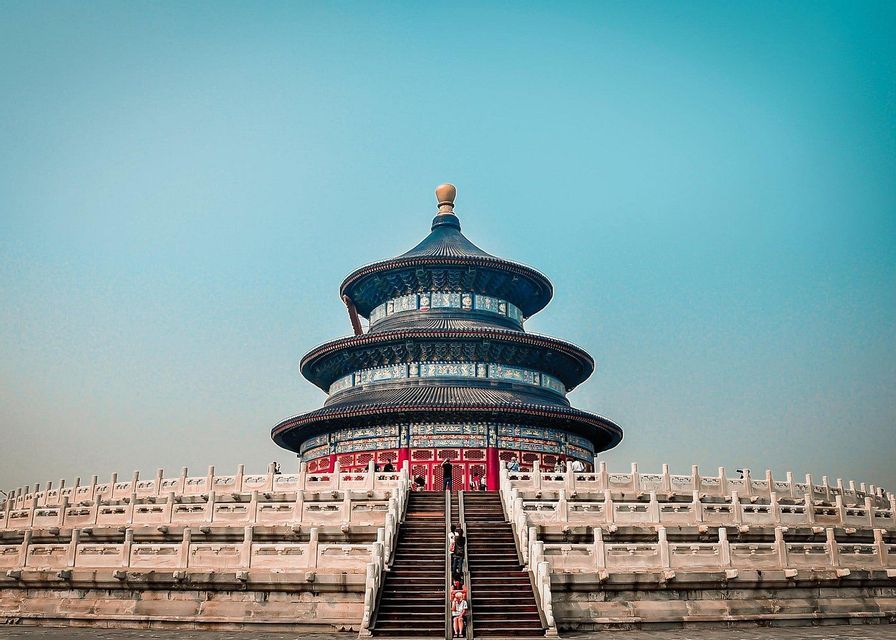 A low-angle view of a traditional, three-tiered Chinese temple with a blue roof, standing atop a wide stone staircase under a clear blue sky.