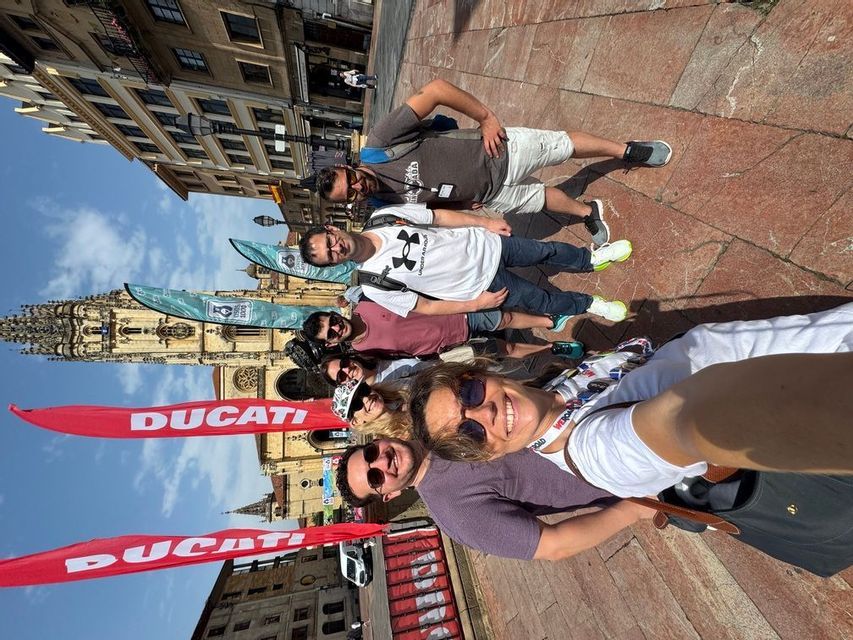 Un grupo de WeRoad sonríe para una selfie en una soleada plaza de la ciudad con una catedral y banderas rojas de Ducati al fondo.