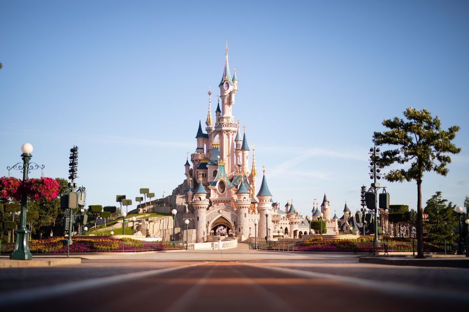 Disneyland Paris Weekend Una prospettiva dal basso di un imponente castello da fiaba rosa e blu, con molteplici torri, situato in fondo a una piazza sotto un cielo sereno.
