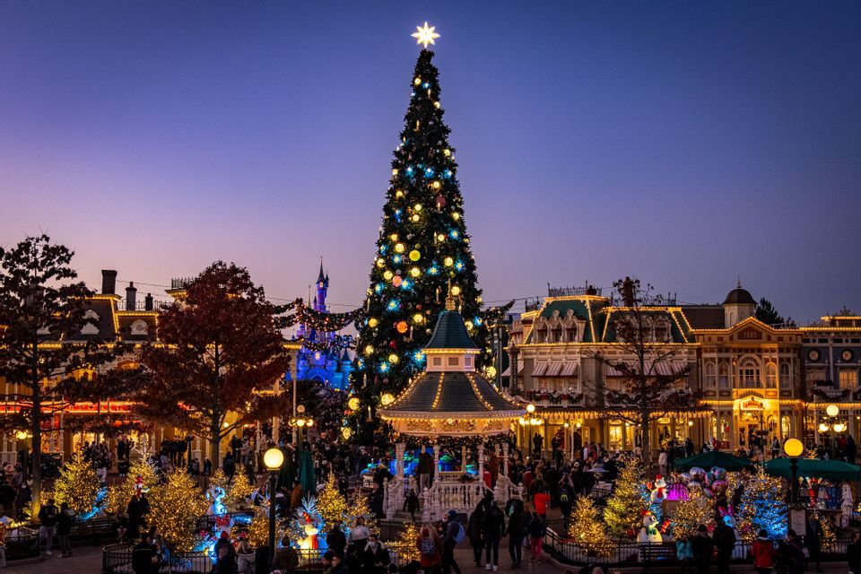 Disneyland Paris Weekend Un grande albero di Natale illuminato con una stella in cima si erge in una piazza affollata decorata con luci festive al tramonto.