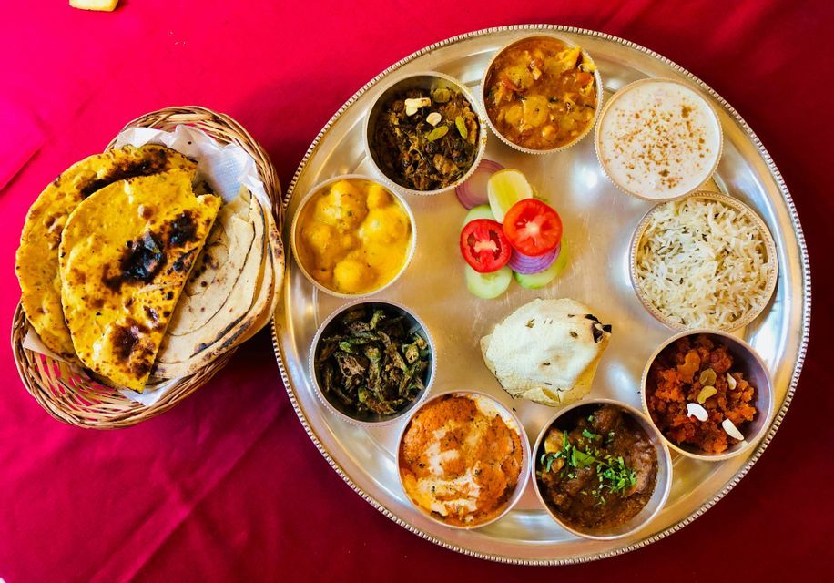 Veduta dall'alto di un thali indiano con diversi curry e riso, accompagnato da un cestino di pane piatto su un panno rosso.