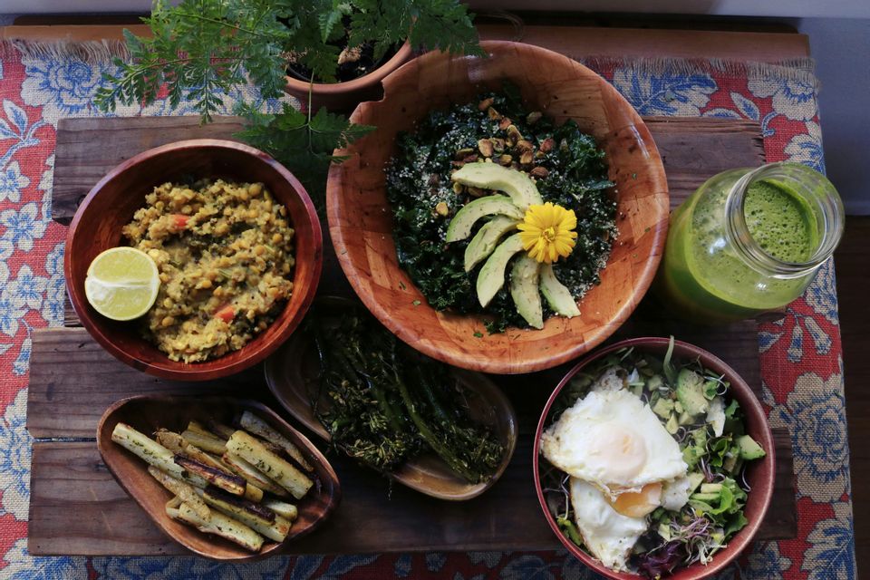 Una veduta dall'alto di un pasto sano e vario, servito in ciotole di legno, che include insalate, verdure arrostite, lenticchie e un frullato verde.