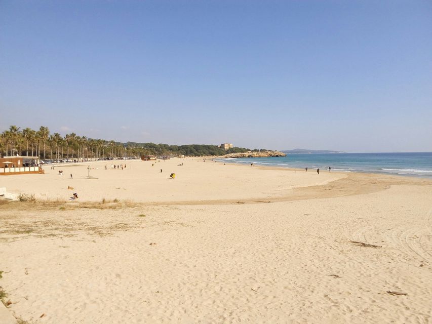 Una spiaggia ampia e sabbiosa con persone sparse lungo la riva, bagnata da acque calme e blu sotto un cielo sereno.