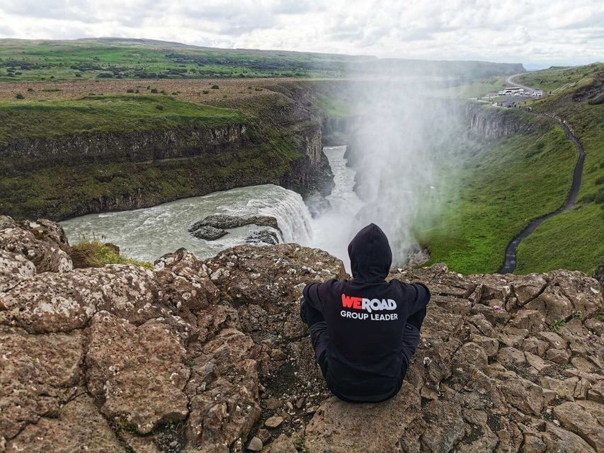 Un accompagnatore WeRoad con una felpa nera è seduto sul bordo di una scogliera rocciosa, affacciato su una potente cascata in un canyon verde.