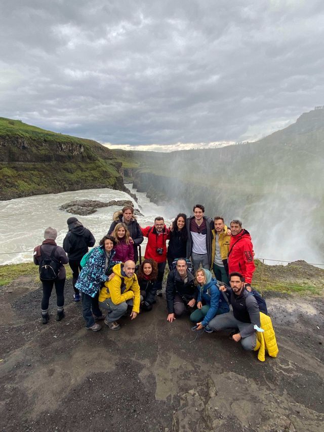 Un voyage en groupe WeRoad posant ensemble au bord d'une falaise, à côté d'une grande cascade brumeuse, sous un ciel nuageux.
