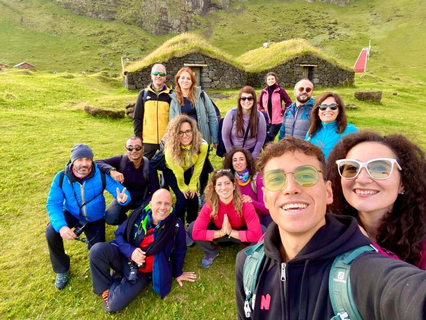 Un groupe WeRoad prend un selfie souriant sur une colline verdoyante, avec des maisons traditionnelles aux toits de tourbe en arrière-plan.