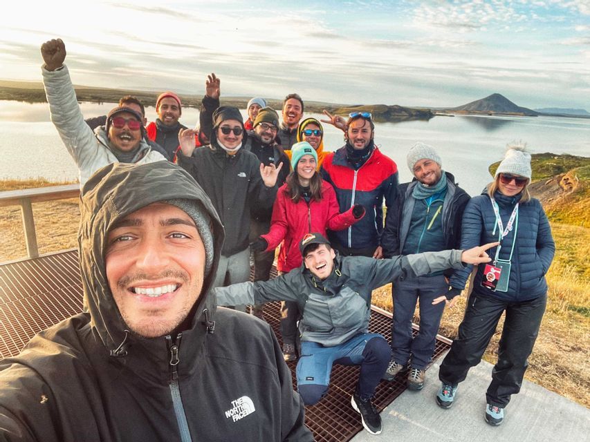 Un grupo de WeRoad sonríe y posa para una selfie grupal en una plataforma de observación con vistas a un lago y montañas al atardecer.