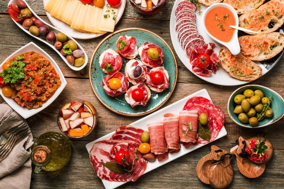 An overhead view of a variety of Spanish tapas, including cured meats, cheeses, olives, and toasts, arranged on a rustic wooden table.