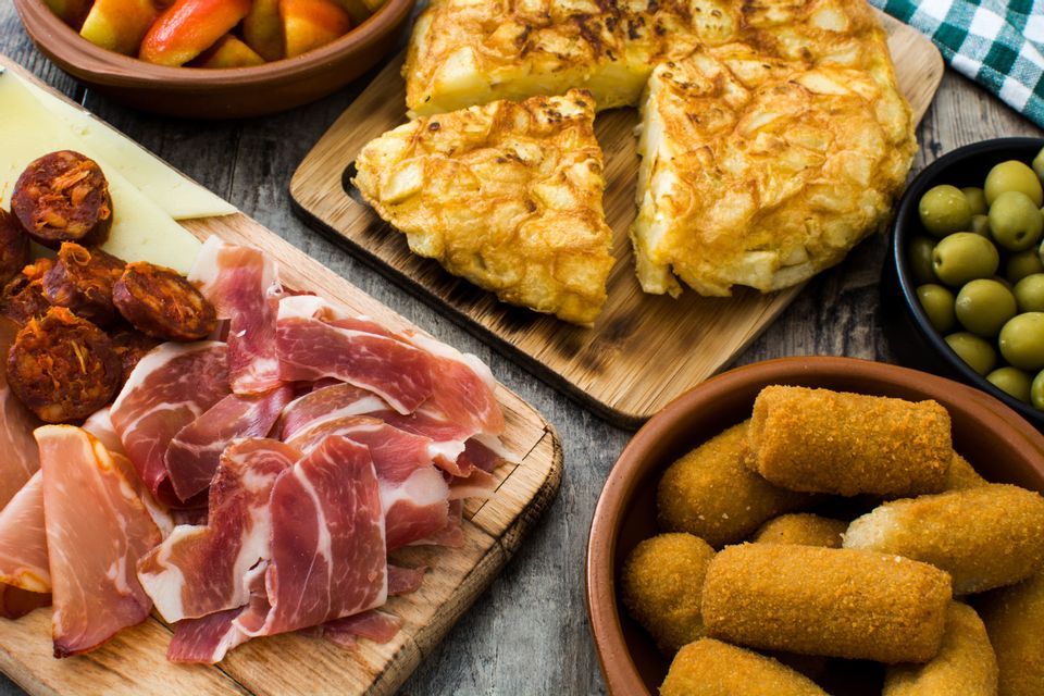 An assortment of traditional Spanish tapas, including tortilla, jamón, croquettes, and olives, laid out on a wooden table.