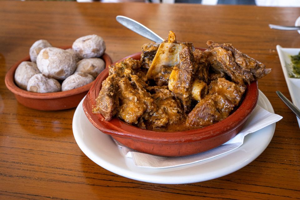 A terracotta bowl of meat stew with bones sits next to a smaller bowl of salted, wrinkled potatoes on a wooden table.