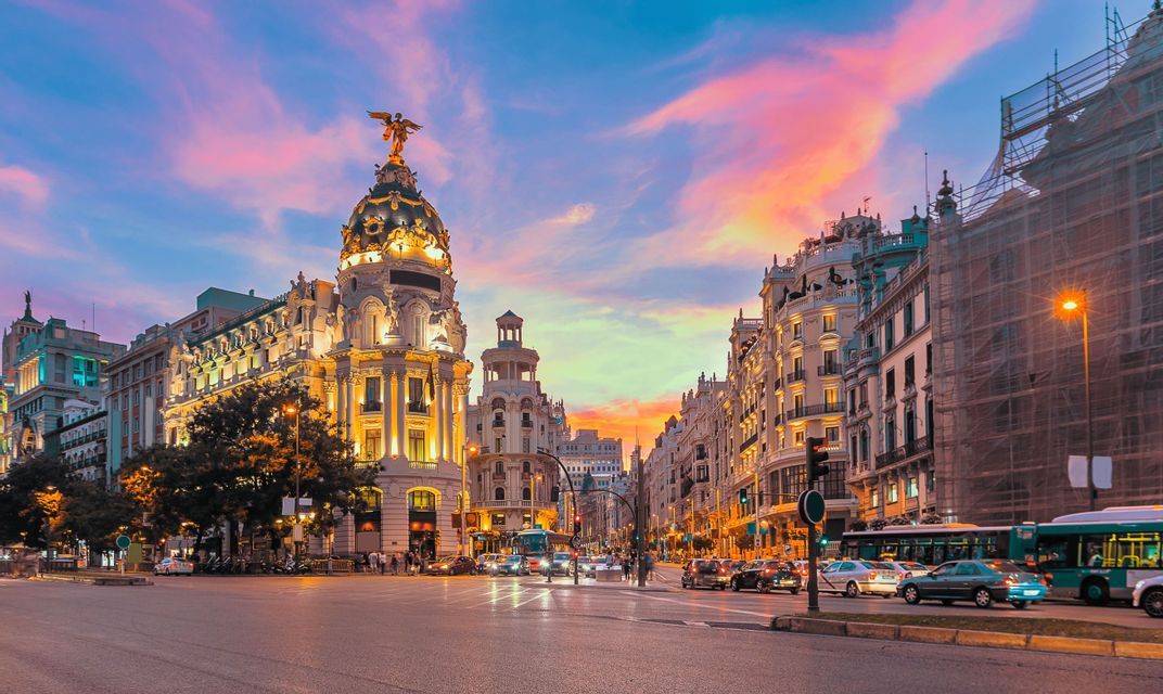 A bustling city street corner with illuminated classical buildings, including one with a domed roof, under a colorful sunset sky.