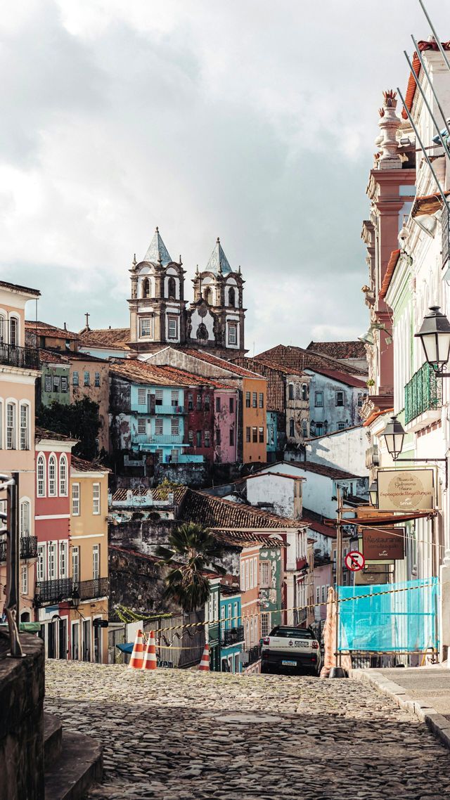 Eine Kopfsteinpflasterstraße führt abwärts durch eine Stadt mit farbenfrohen, historischen Gebäuden und einer im Hintergrund sichtbaren Kirche.