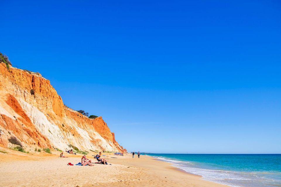 People relax on a sandy beach at the base of tall, reddish-orange cliffs next to the turquoise sea under a clear blue sky.