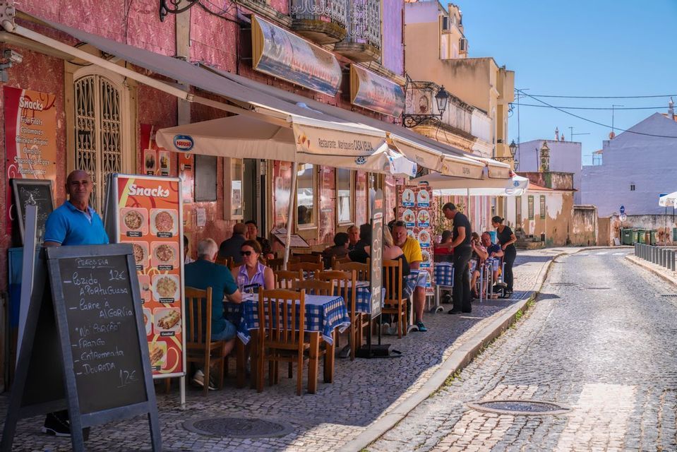 People dine at tables with blue checkered tablecloths outside a pink restaurant on a sunny cobblestone street.