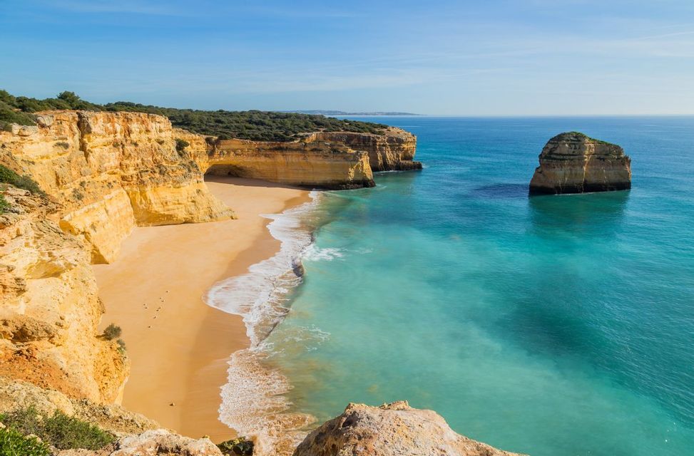 A high-angle view of a secluded sandy beach with turquoise water, framed by golden limestone cliffs under a clear blue sky.