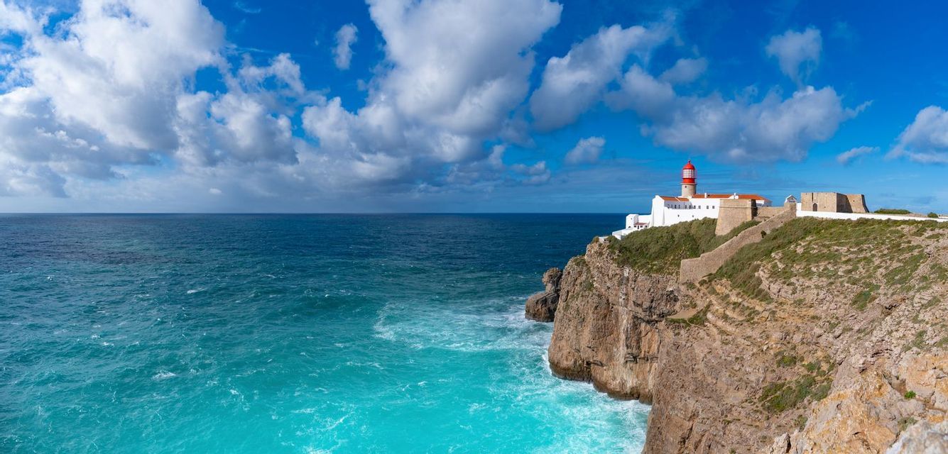 A white lighthouse with a red top stands on a rocky cliff overlooking the turquoise ocean under a partly cloudy sky.