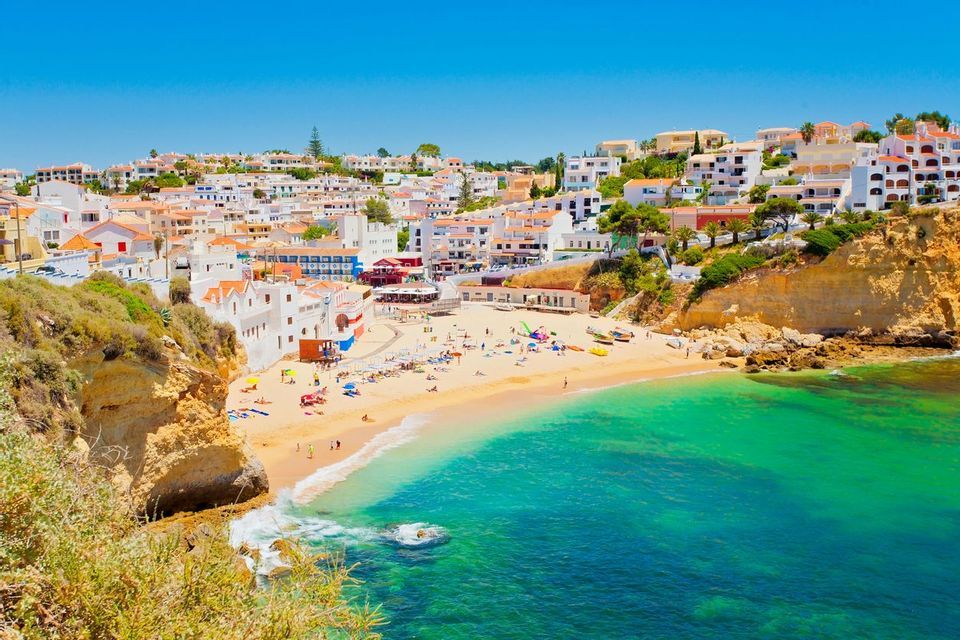 A coastal town with white buildings overlooks a sandy beach cove filled with sunbathers and turquoise water under a clear blue sky.