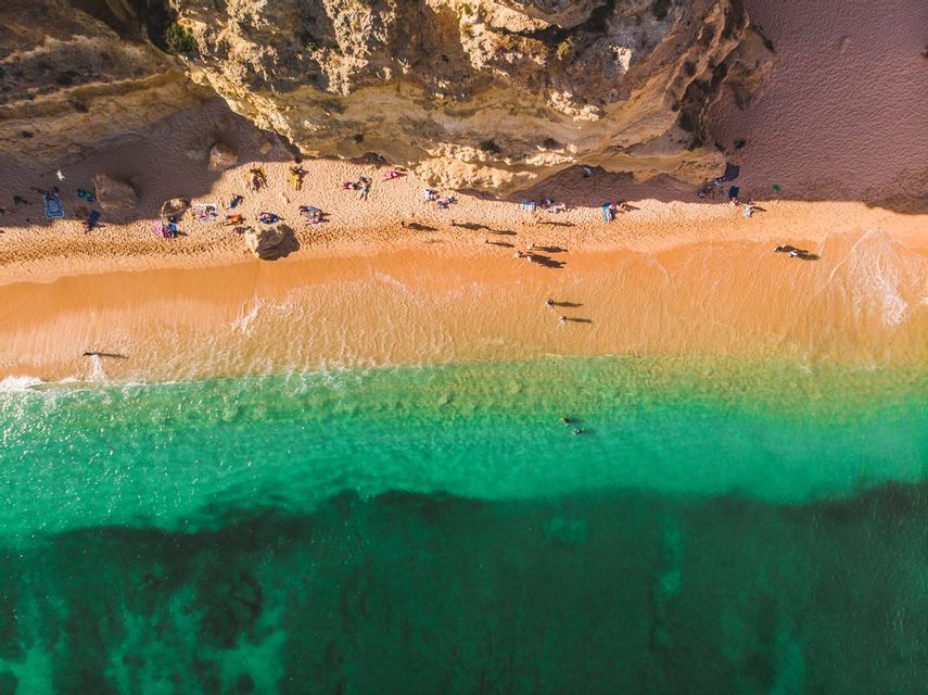 An aerial top-down view of people on a golden beach next to a rocky cliff and turquoise water.