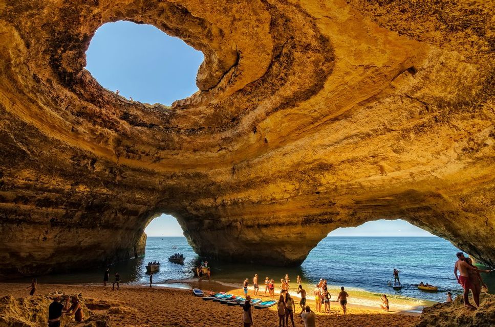 A WeRoad group trip gathers on a sandy beach inside a large sea cave with an oculus in the ceiling revealing the blue sky.