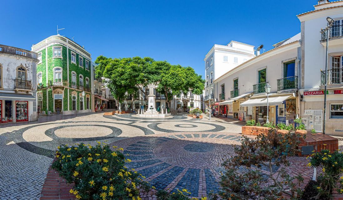 A sunlit European town square with patterned cobblestone streets, surrounded by traditional buildings, including one with green tiles.