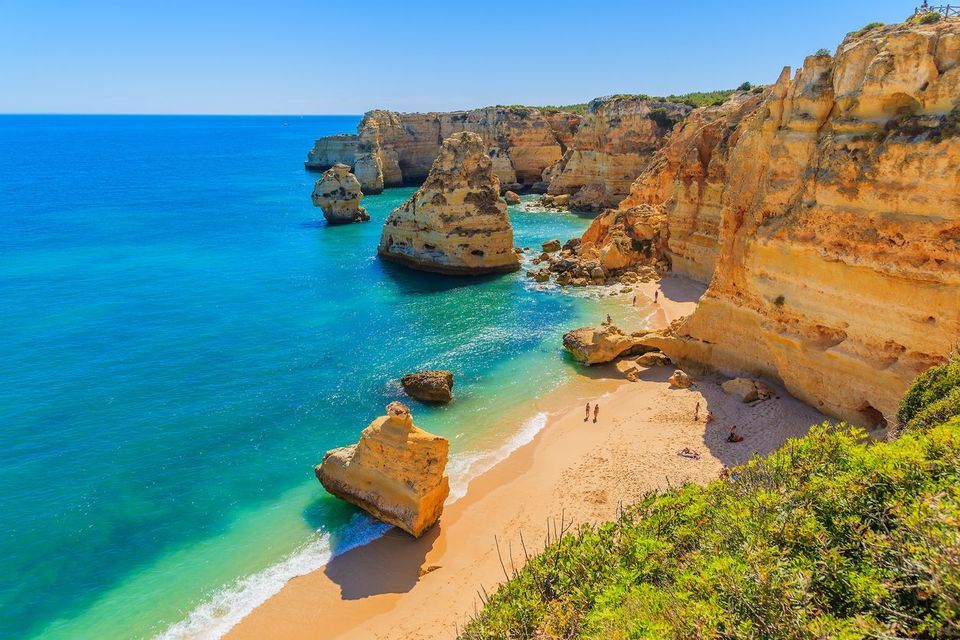 Tall golden cliffs and sea stacks line a sandy beach with turquoise water under a clear blue sky.