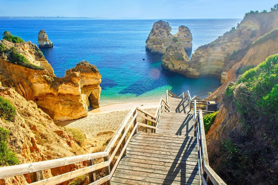 Wooden stairs descend a rocky cliff to a secluded sandy beach with turquoise water and large rock formations.