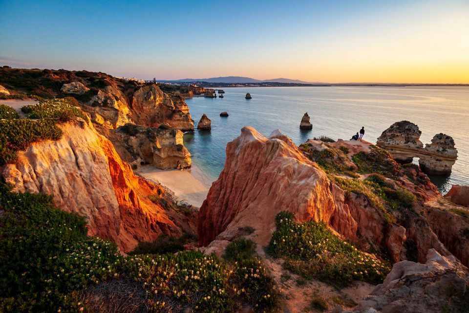 Rugged red cliffs covered in green vegetation overlook a calm sea with sea stacks and a sandy cove at sunset.
