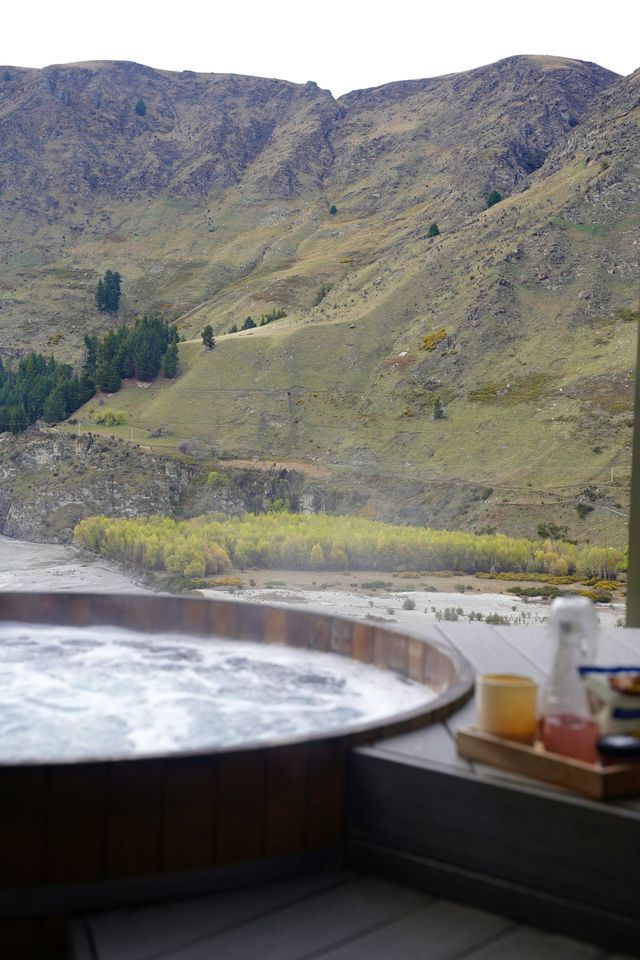 Una tinozza idromassaggio in legno con acqua gorgogliante su una terrazza, con vista su una valle fluviale e un fianco di montagna erboso.
