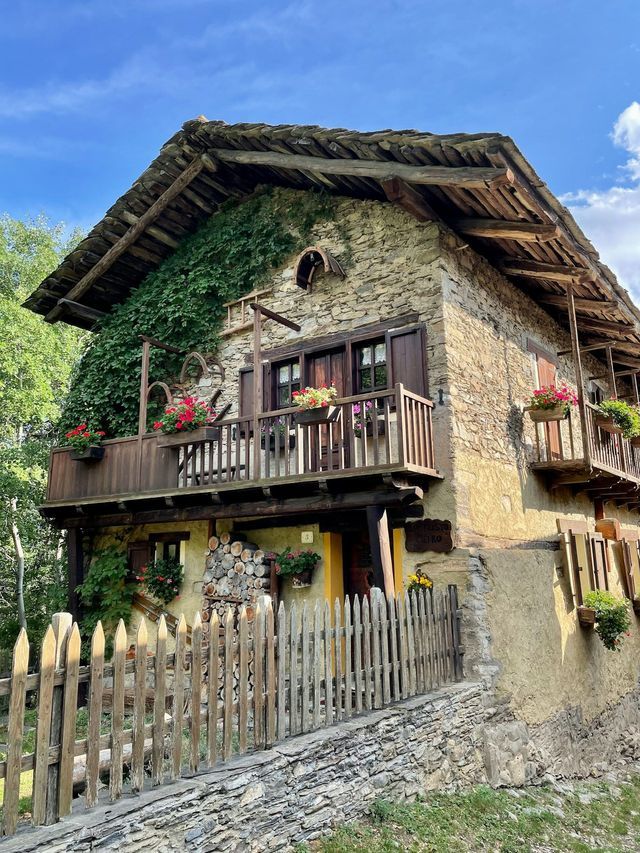 Una casa rustica in pietra con muri coperti di edera, balconi in legno fioriti e una staccionata in un ambiente rurale.