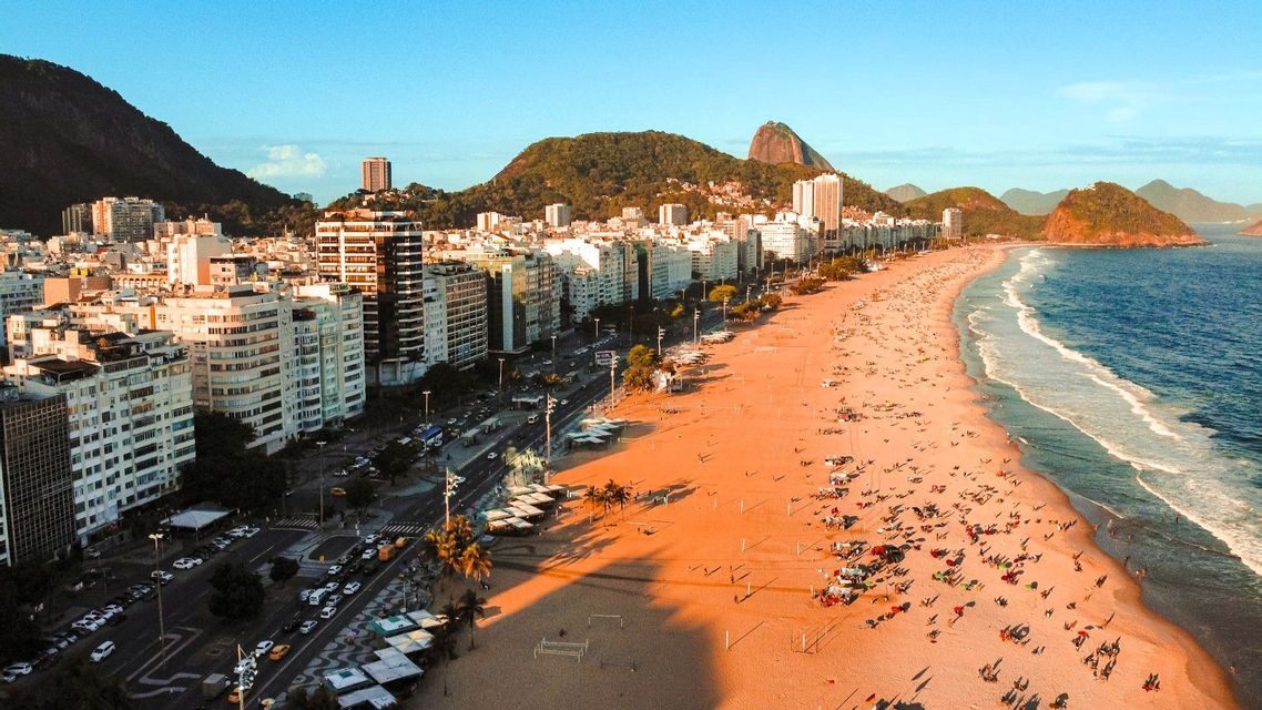 Eine Luftaufnahme eines belebten Stadtstrandes bei Sonnenuntergang, mit Wellen, die an den Sand rollen, und Bergen, die sich hinter den Gebäuden erheben.