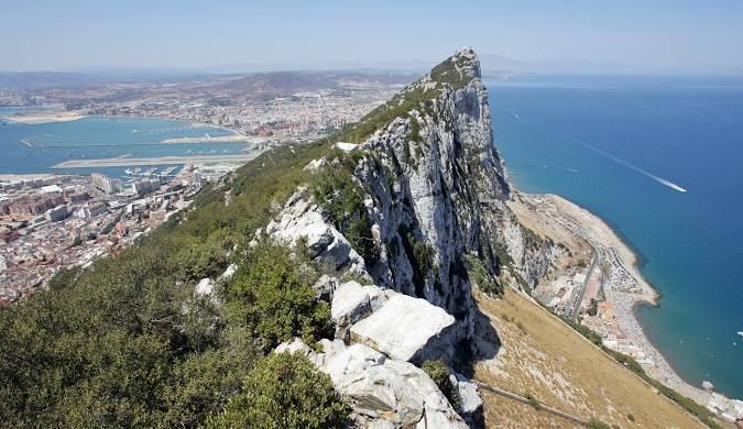 Una vista elevada desde una cresta rocosa de montaña, dominando una ciudad costera, un puerto y el mar azul profundo en un día soleado.