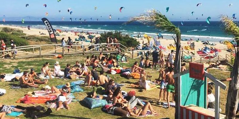 Un gran grupo de personas tomando el sol en un césped con vistas a una playa llena de kitesurfistas bajo un cielo azul claro.