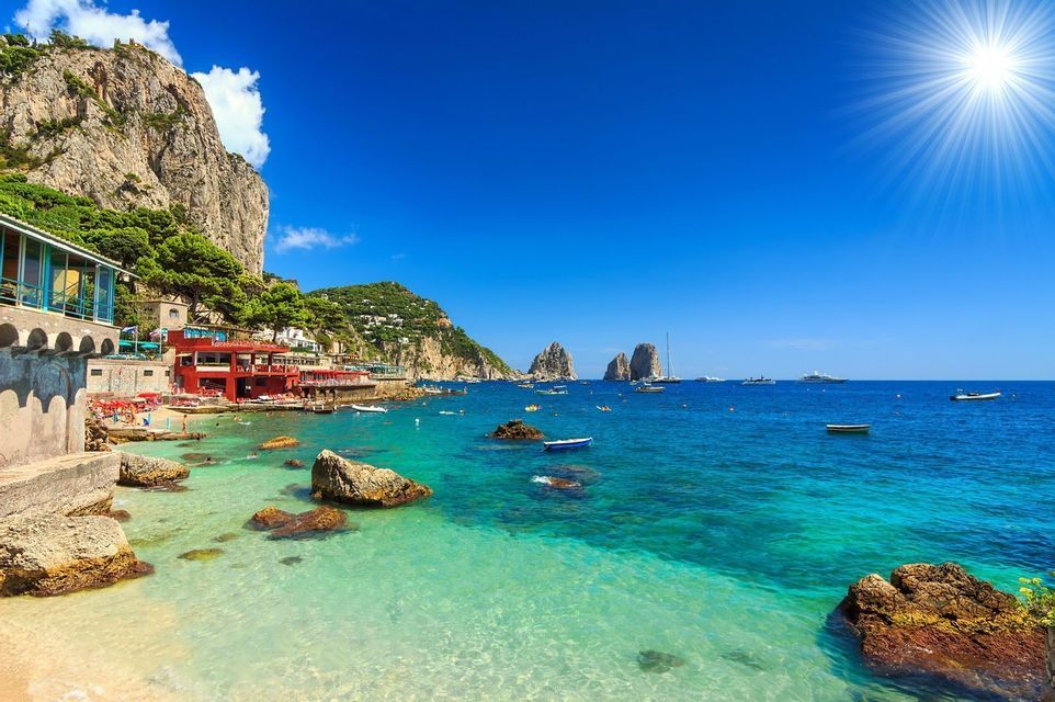 A cliffside village overlooks a turquoise bay with swimmers, boats, and large rock stacks under a bright, sunny sky.