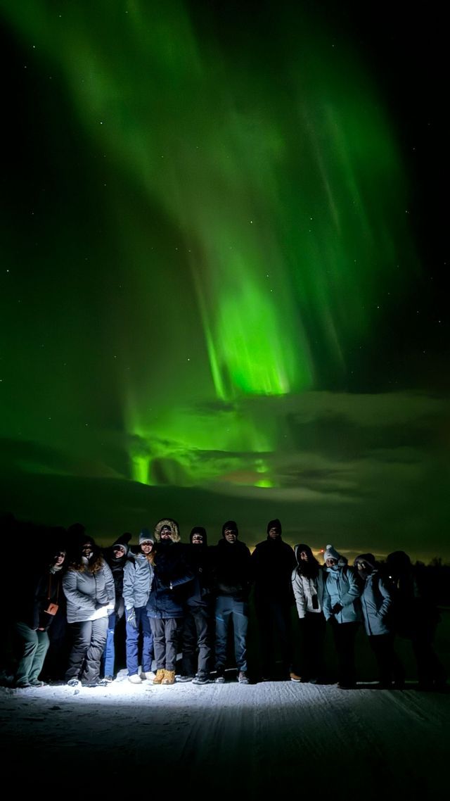 Un viaggio di gruppo WeRoad in un paesaggio innevato di notte, in posa per una foto sotto il bagliore verde dell'Aurora Boreale.