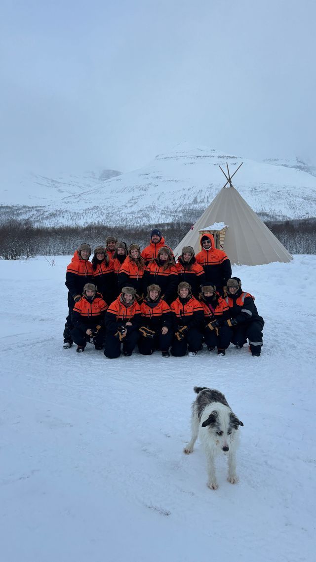 Un viaggio di gruppo WeRoad con abbigliamento invernale coordinato posa in un paesaggio innevato, con un cane in primo piano, un teepee e montagne sullo sfondo.