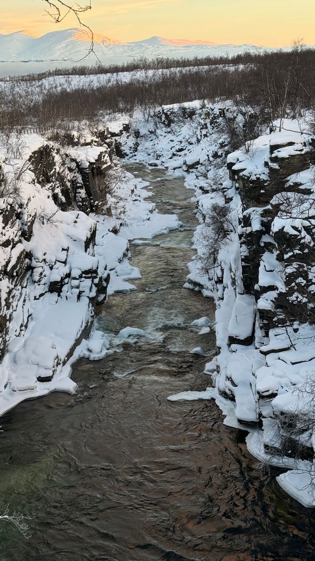 Une rivière coule à travers un canyon profond et enneigé, flanqué de falaises rocheuses, avec des montagnes enneigées visibles au loin.
