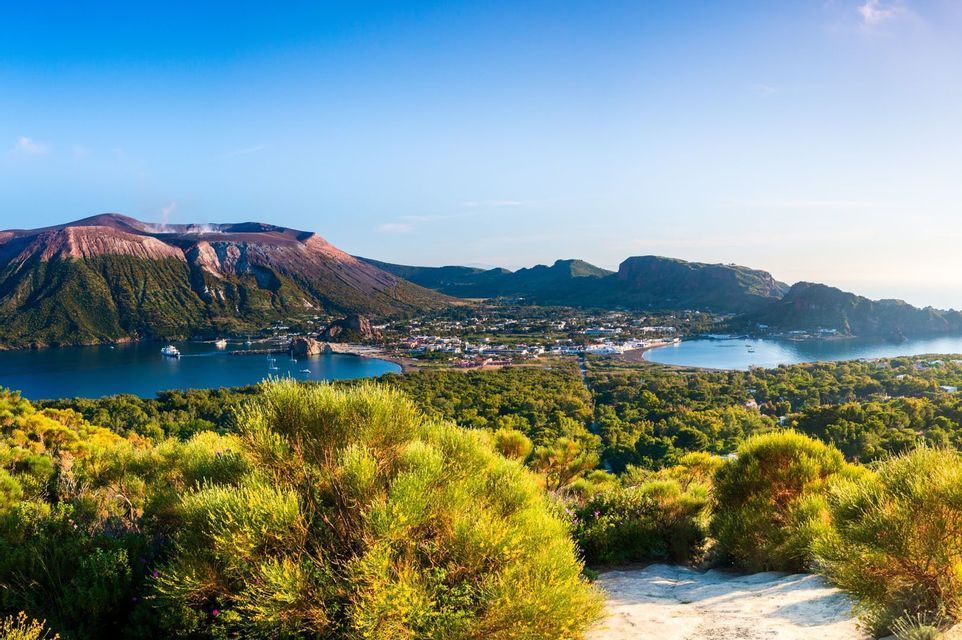 Vista dall'alto di un'isola vulcanica che mostra una città costiera, una baia blu con barche e il cratere del vulcano sotto un cielo limpido.