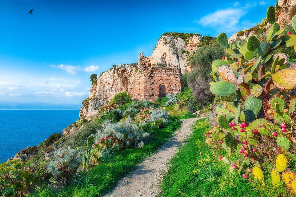 Un sentiero sterrato si snoda tra fichi d'India su una scogliera, conducendo a un rudere in pietra affacciato sul mare.