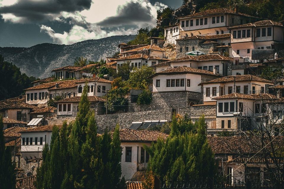 Un village historique de maisons blanches aux murs de pierre et aux toits de tuiles est niché à flanc de colline verdoyante sous un ciel nuageux.