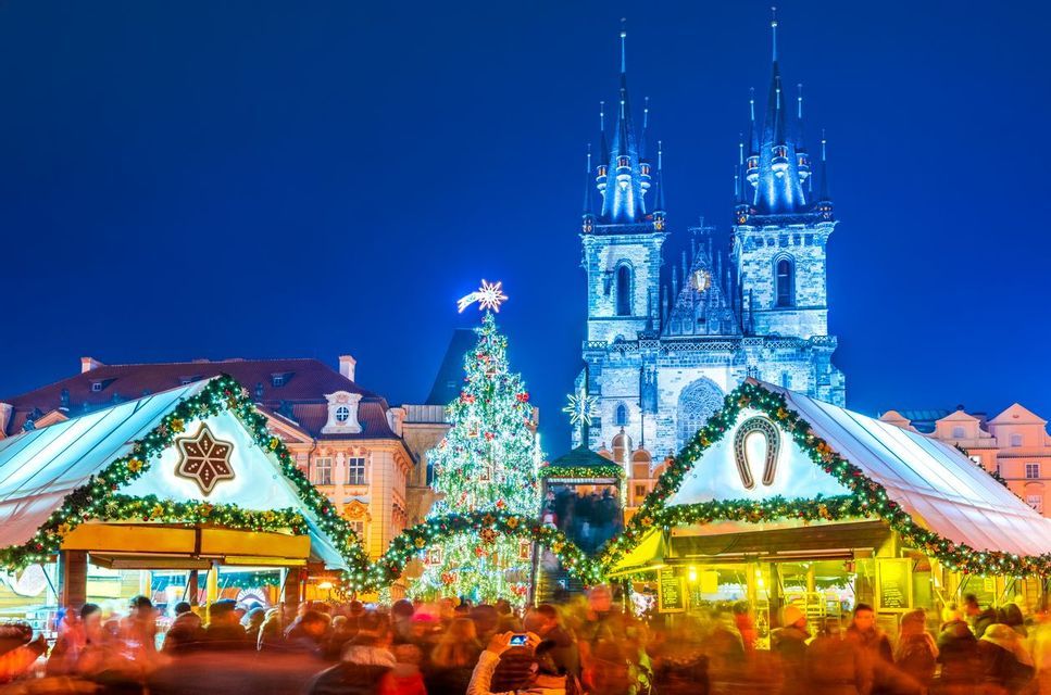 A crowded Christmas market at night with an illuminated tree and stalls in front of a large Gothic church.