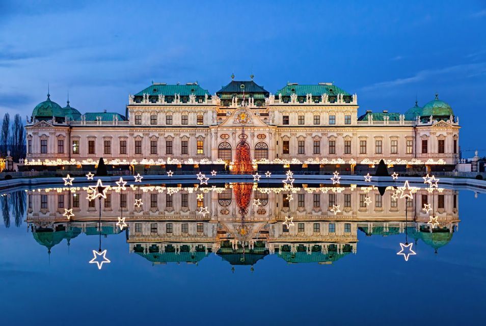 An ornate palace illuminated for Christmas with a large lighted tree and stars, reflected perfectly in a pool of water at dusk.