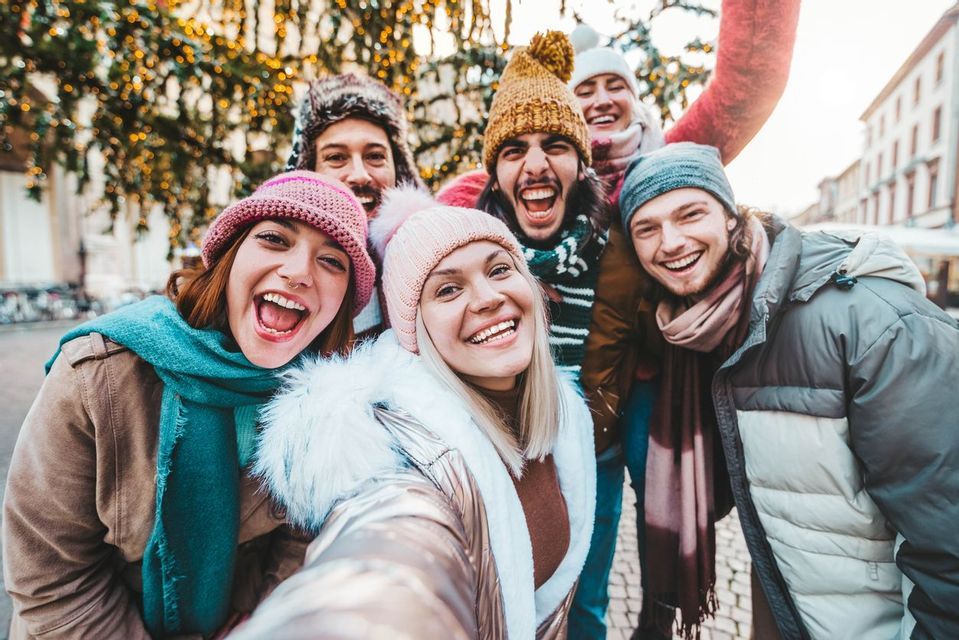 A WeRoad group trip of smiling friends in winter clothes taking a selfie together outdoors with festive lights.