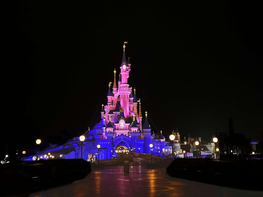 Ein großes Schloss, beleuchtet mit rosa und blauen Lichtern vor einem dunklen Nachthimmel, während eine Person auf dem nassen Weg darauf zugeht.