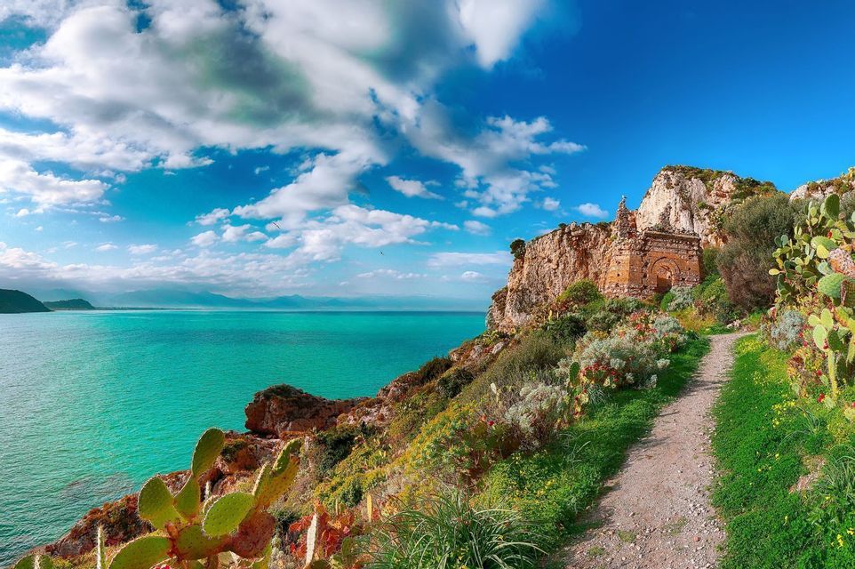 Un sentiero sterrato conduce a un monumento in pietra su una scogliera verde a picco su un mare turchese sotto un cielo nuvoloso.