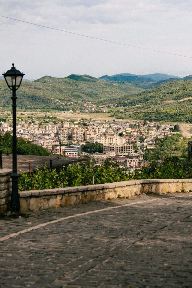 Una ciudad enclavada en un valle verde, vista desde un camino empedrado alto con una farola negra en primer plano.